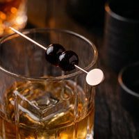 A stainless cocktail pick with two cherries, resting across a rocks glass of whisky in a dark bar